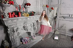 A beautiful pensive girl in a white sweater and pink skirt by the fireplace