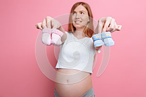 Beautiful pensive pregnant woman holding blue and pink baby booties, posing in the studio on a pink background