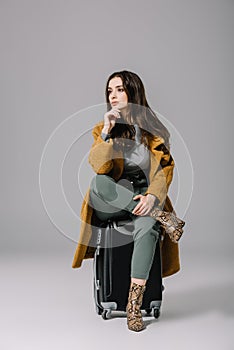 beautiful pensive woman in beige coat sitting on travel bag on grey.