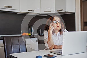 Beautiful pensive woman pensioner in glasses sitting at home at the table using a laptop