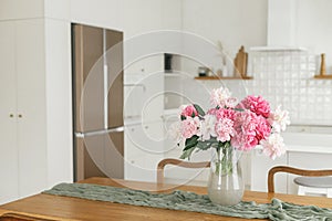 Beautiful peonies in vase on wooden table on background of stylish white kitchen with appliances in new scandinavian house. Modern