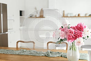 Beautiful peonies in vase on wooden table on background of stylish white kitchen with appliances in new scandinavian house. Modern