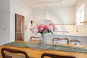 Beautiful peonies in vase on wooden table on background of stylish white kitchen with island, wooden shelves and appliances in new