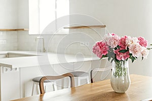 Beautiful peonies in vase on wooden table on background of stylish white kitchen with island, wooden shelves and appliances in new