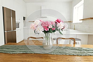 Beautiful peonies in vase on wooden table on background of stylish white kitchen with island, wooden shelves and appliances in new