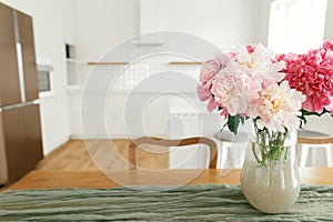 Beautiful peonies in vase on wooden table on background of stylish white kitchen with island, wooden shelves and appliances in new