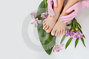 Beautiful perfect skin female legs feet top view with tropical flowers and green palm leaf. Hands of beautician in pink gloves.