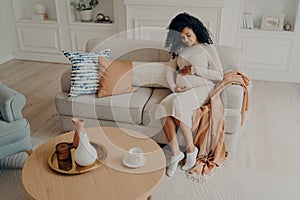 Beautiful photo afro american lady sitting on cozy sofa and expecting baby, looking down on her belly