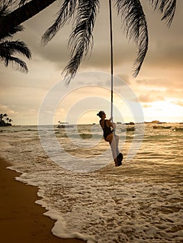 Beautiful image of sexy yougn woman in swimsuit swinging on the rope tied to the palm tree over the ocean beach