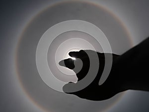 Beautiful photograph of the sun with a circular rainbow surrounded by a bright sky and white clouds with shadows of hands reaching