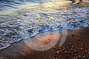 A beautiful picture of a sea wave with foam running onto the beach in the rays of the setting sun. Sea waves at sunset