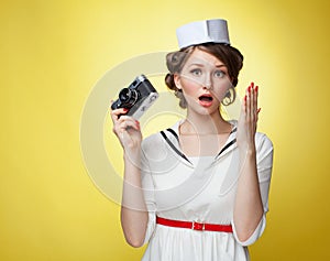 Beautiful pin-up girl dressed a sailor holding a vintage camera and screams behind his hand. Yellow background, close up