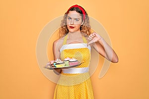 Beautiful pin-up woman with blue eyes holding chocolate cupcakes over yellow background with angry face, negative sign showing