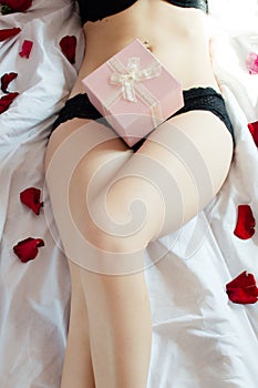 Beautiful pink present lying on adorable female stomach legs up in white bed surrounded by rose flower petals