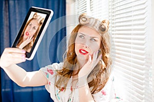 Beautiful pinup girl happy smiling and taking selfie or selfy photo with tablet pc at home by window blinds closeup portrait