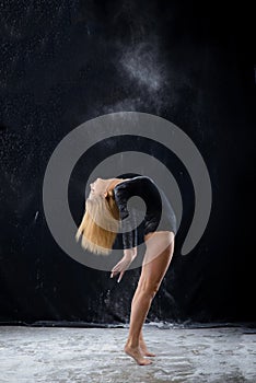 Beautiful plump blonde girl wearing a black gymnastic bodysuit covered with clouds of the flying white powder jumps dancing