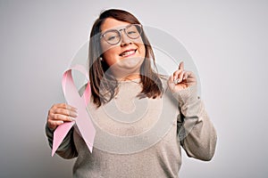 Beautiful plus size woman holding breast cancer awareness pink ribbon over isolated background surprised with an idea or question