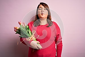 Beautiful plus size woman holding romantic bouquet of natural tulips flowers over pink background making fish face with lips,