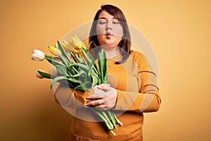 Beautiful plus size woman holding romantic bouquet of natural tulips flowers over yellow background making fish face with lips,