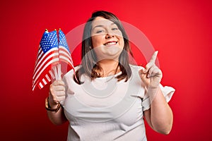 Beautiful plus size woman holding united states of america flag over red background surprised with an idea or question pointing