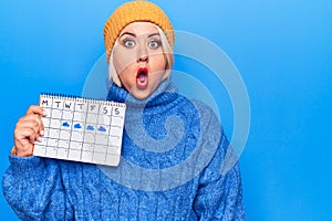 Beautiful plus size woman holding weather calendar showing rainy week over blue background scared and amazed with open mouth for