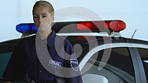 Beautiful police woman standing near cop car and looking into camera, on duty