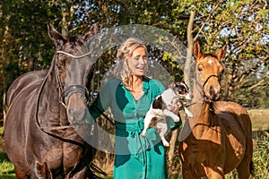 Beautiful portrait of a blond smiling girl with her horse, foal and dog in the forest. Wearing a green dress