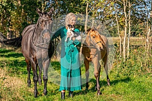 Beautiful portrait of a blond smiling girl with her horse, foal and dog in the forest. Wearing a green dress