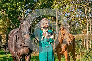 Beautiful portrait of a blond smiling girl with her horse, foal and dog in the forest. Wearing a green dress