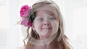 beautiful portrait blonde of a girl child in a pink dress and with rose flower in her head background of the window