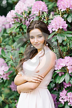 Beautiful portrait of a lovely girl who stands surrounded by lilac pink flowers blooming rhododendron in the spring in the garden.