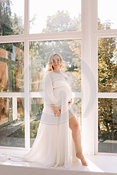 Beautiful portrait of pregnant woman in white dress. Concept of perfect pregnancy. Woman put her hand on belly. Future