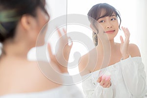 Beautiful portrait young asian woman applying cream moisturizer or lotion skin care cosmetic on face looking mirror