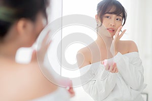 Beautiful portrait young asian woman applying cream moisturizer or lotion skin care cosmetic on face looking mirror