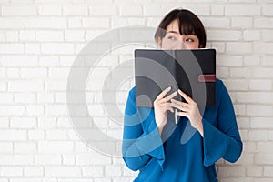 Beautiful portrait young asian woman happy hiding behind covering the book with cement or brick concrete background