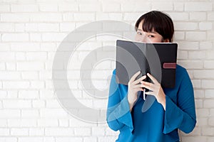 Beautiful portrait young asian woman happy hiding behind covering the book with cement or brick concrete background