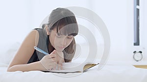 Beautiful portrait young asian woman relax lying on bed thinking idea and writing book or diary in the bedroom at home