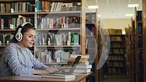 Beautiful positive caucasian female student with big headphones working at table in spacious library in front of laptop