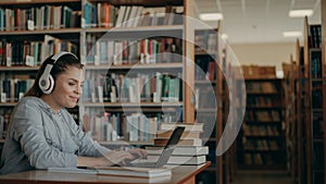 Beautiful positive caucasian female student with big headphones working at table in spacious library in front of laptop