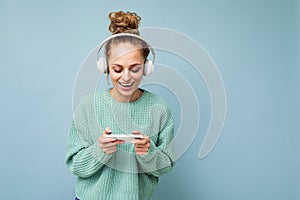 Beautiful positive smiling young woman wearing blue sweater isolated over blue background wearing white bluetooth