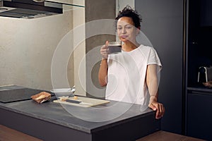 Beautiful positive woman enjoys her morning coffee, smiling looking at camera, standing at the kitchen table