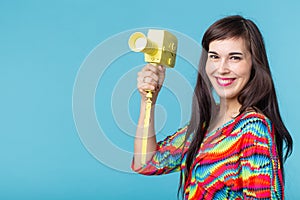 Beautiful positive young brunette woman holding in her hands a model of a yellow camcorder posing on a blue background