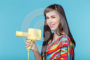 Beautiful positive young brunette woman holding in her hands a model of a yellow camcorder posing on a blue background