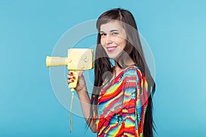Beautiful positive young brunette woman holding in her hands a model of a yellow camcorder posing on a blue background