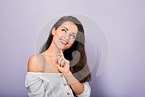 Beautiful positive young casual woman with hand under the face thinking and looking up in white shirt on purple background