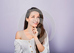Beautiful positive young casual woman with hand under the face thinking and looking up in white shirt on purple background