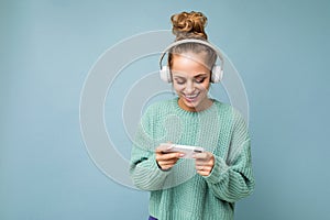 Beautiful positive young woman wearing blue sweater isolated over blue background wearing white bluetooth wireless