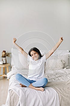 Beautiful pre-teen girl in casual clothes do stretching exercises seated on bed in cozy bedroom in the morning