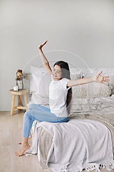 Beautiful pre-teen girl in casual clothes do stretching exercises seated on bed in cozy bedroom in the morning