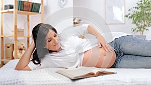 Beautiful pregnant 40 years old woman lying in bedroom and stroking her big belly when reading book
